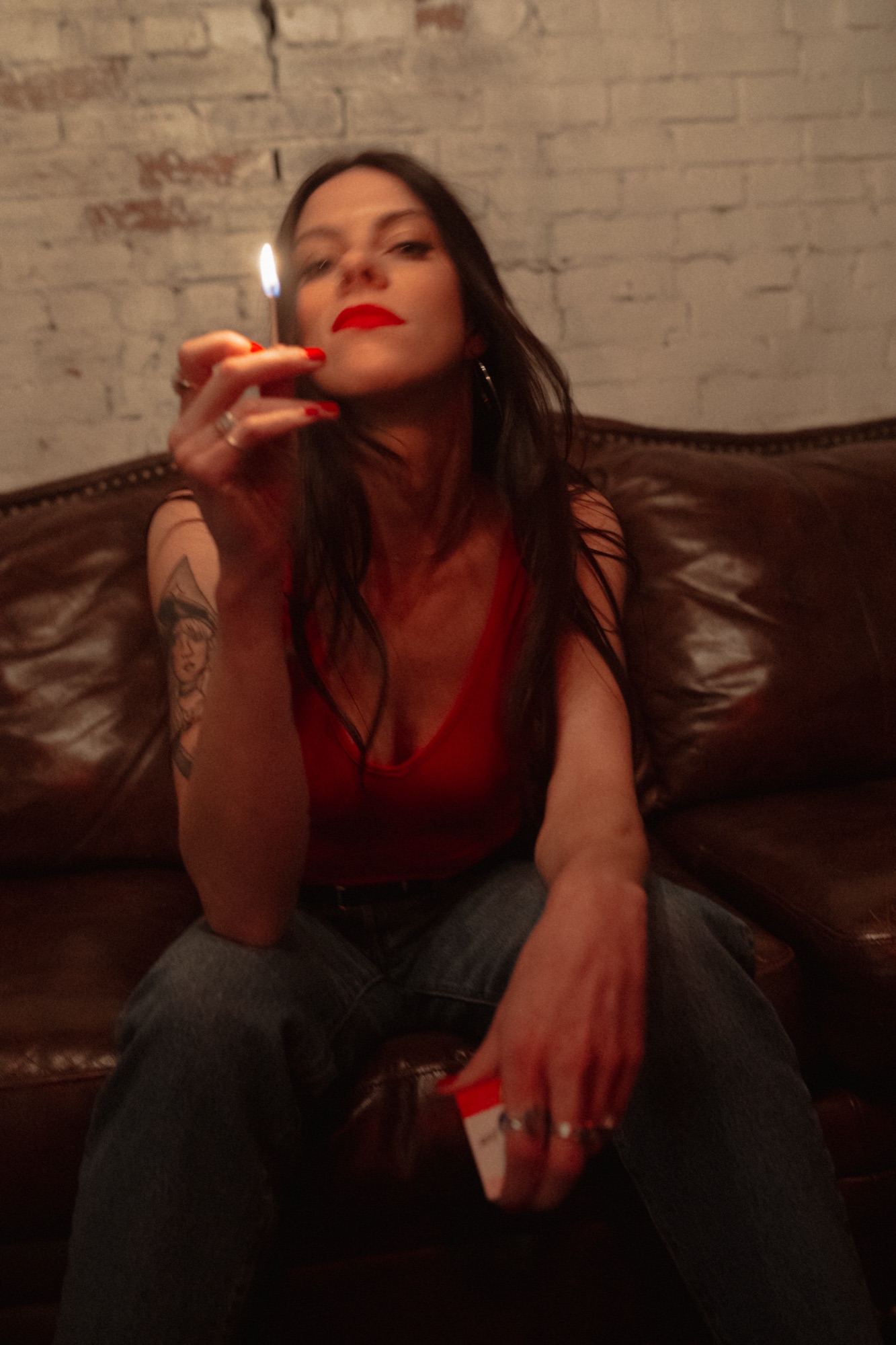 Elyse Aeryn seated on a leather couch in a red top, holding a lit match in front of her face